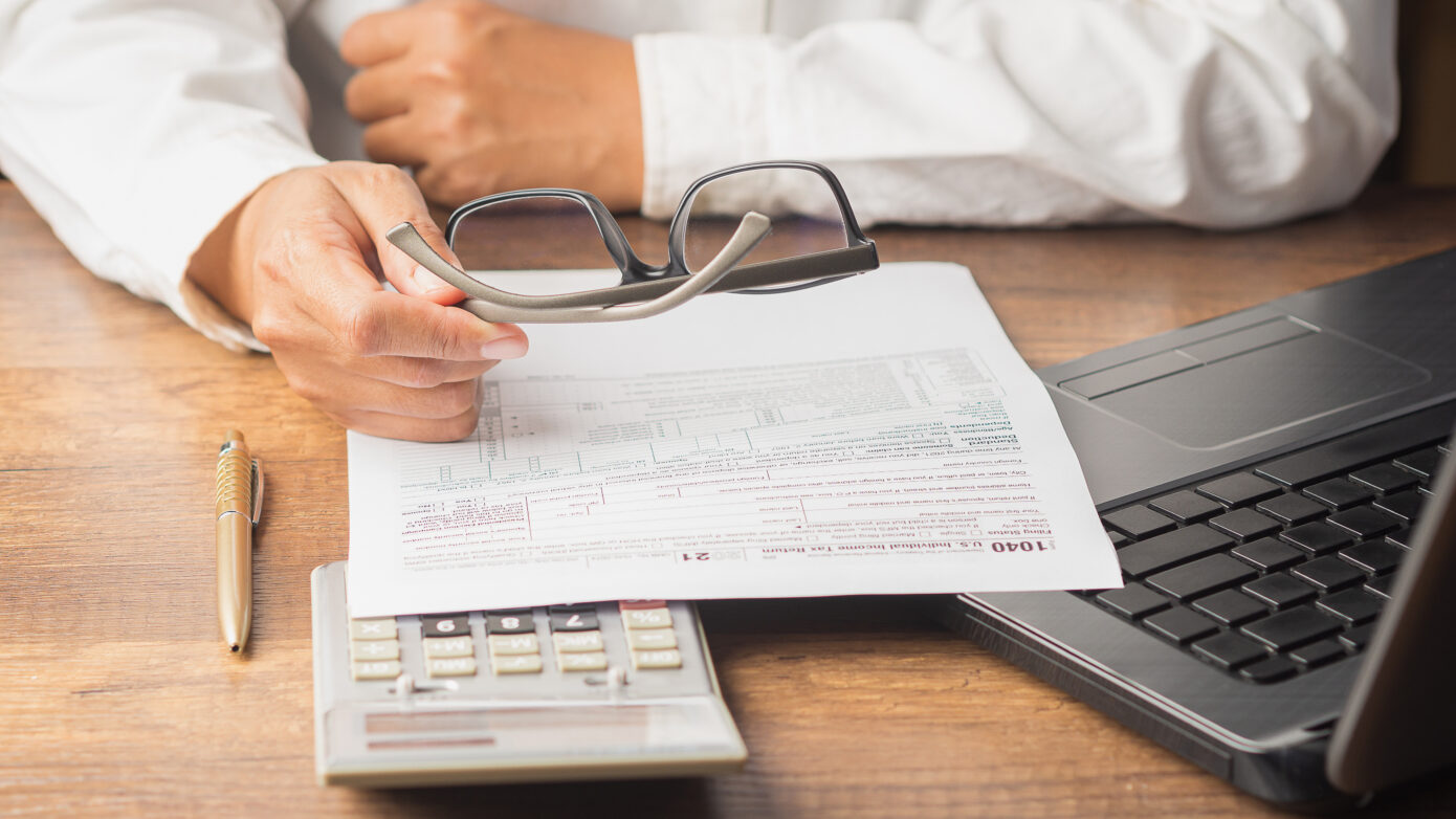 Businessman in a white shirt holds his glasses. A payroll tax form is in front of him on the table accompanied by a calculator and a yellow pen.