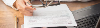 Businessman in a white shirt holds his glasses. A payroll tax form is in front of him on the table accompanied by a calculator and a yellow pen.