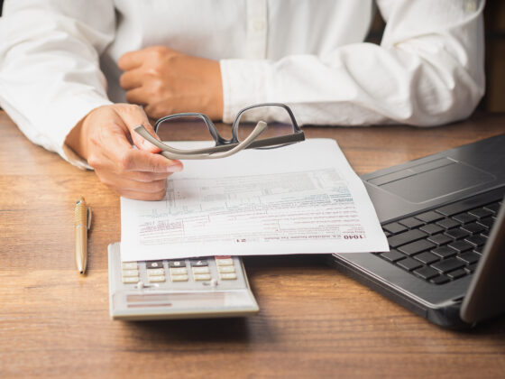 Businessman in a white shirt holds his glasses. A payroll tax form is in front of him on the table accompanied by a calculator and a yellow pen.