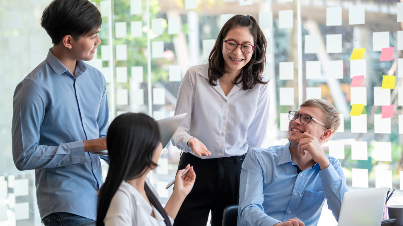 Group of junior corporate office workers gather around a laptop. They are enjoying their conversation.