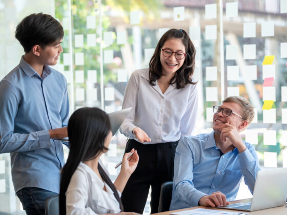 Group of junior corporate office workers gather around a laptop. They are enjoying their conversation.