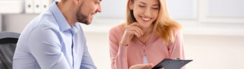 Two accountants, a white young man wearing a blue shirt and a white young woman wearing a pink cardigan, discuss payments and look at a black plastic clipboard.