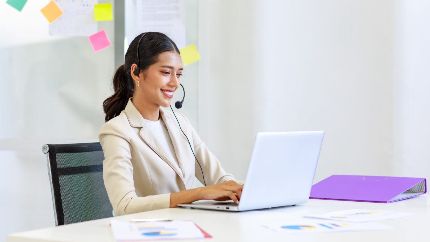 Young Asian professional woman speaking through a headset while using a silver laptop.