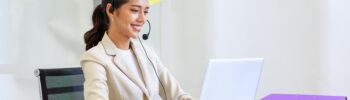 Young Asian professional woman speaking through a headset while using a silver laptop.